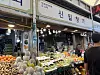 Fruit and vegetables fill a brightly lit market stall in Seoul, with Korean signage above and shoppers nearby.