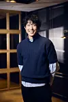 Song In-hyeok smiling in a warmly lit interior while wearing a dark sweater.