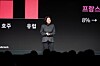 Presenter on stage beside a screen displaying red bars, regional labels and an 8% figure.