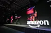 Amazon logo on a dark event stage beside colorful presentation screens and a distant presenter.