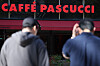 Two people stand outside a Caffè Pascucci storefront beneath the red café sign.