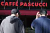 Two people stand outside a Caffè Pascucci storefront beneath the red café sign.