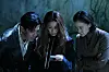 Three people look at a small object together in a dark wooded setting.